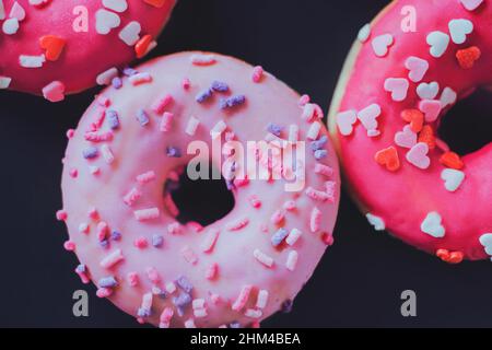 On a dark background there are delicious sweet pink donuts with multicolored sprinkles in the form of hearts. A nice delicious gift for Valentine's da Stock Photo