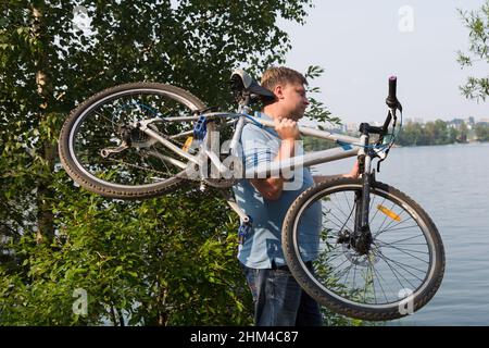 A frustrated adult man carries a broken bicycle on his shoulder. Stock Photo