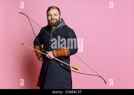Studio shot of young smiling bearded man in image of medieval warrior, archery in chain armor isolated over pink studio background Stock Photo