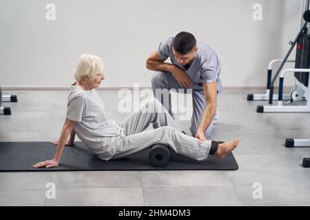 Aged lady sitting on mat with legs on roll Stock Photo