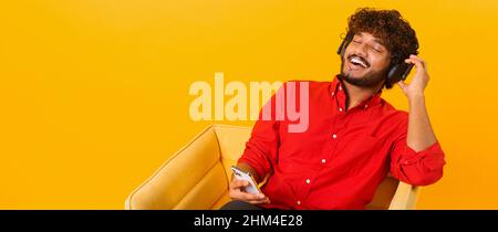 Joyful middle eastern man wearing wireless headphones holding smartphone in hand, enjoying music. Ecstatic cheerful multiracial guy listening favorite tracks, isolated on orange Stock Photo