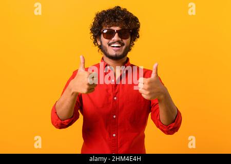 Like, excellent job. Pleased man with beard wearing sunglasses smiling to camera and gesturing thumbs up, approval hand sign, satisfied with result. Indoor studio shot isolated on orange background Stock Photo