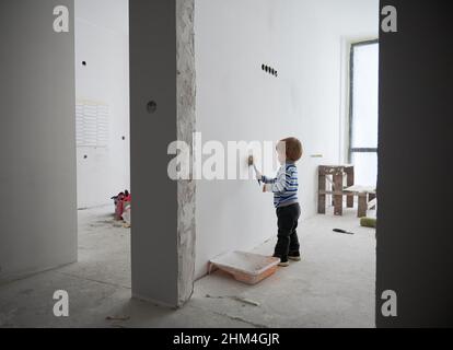 Adorable child painting white wall between two rooms under renovation. Cute baby boy using paint brush while renovating apartment. Concept of home renovation, refurbishment and childhood. Stock Photo