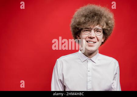 A handsome curly-haired man in a shirt and glasses smiles at the camera on a red background. Unusual hairstyle. The man has poor eyesight. Stock Photo