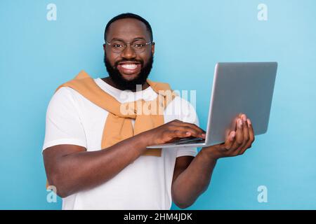Photo of cheerful programmer guy hold netbook typing wear white t-shirt jumper front-tie isolated blue color background Stock Photo