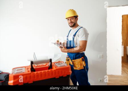 Smiling handyman worker, builder in uniform and hard hat looking at camera, making notes on a clipboard while standing indoors Stock Photo
