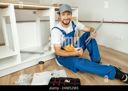 Smiling handyman in overalls holding a part of kitchen cabinet while installing furniture, sitting on the floor Stock Photo