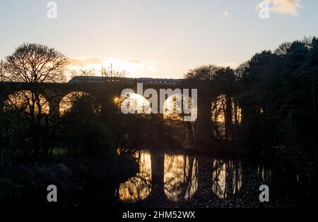 Sun peeks through one arch of a railway viaduct at sunset as a train speeds overhead Stock Photo