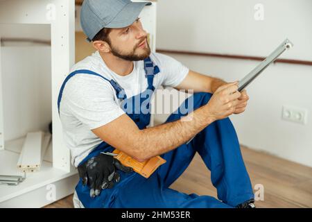 Busy male worker in overalls assembling kitchen furniture, sitting on the floor Stock Photo