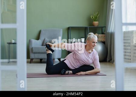 Active senior female practicing yoga asanas on the floor of living-room while sitting on mat and stretching legs and arms Stock Photo