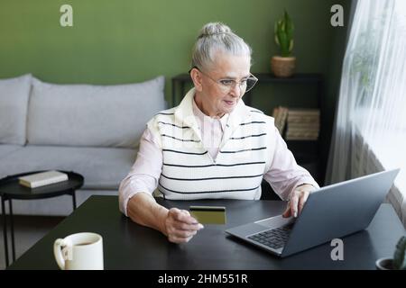 Contemporary senior female in casual clothes looking at laptop screen while making online payment for food products from supermarket Stock Photo