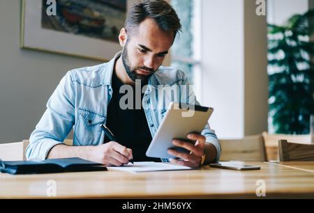 Focused freelancer writing notes and using tablet Stock Photo