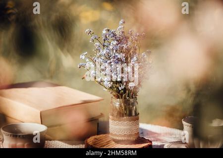 Dream like view through apple blossoms to table, set with tea cups and books. Bouquet of wildflowers Myosotis arvensis forget me nots in handmade jar. Stock Photo