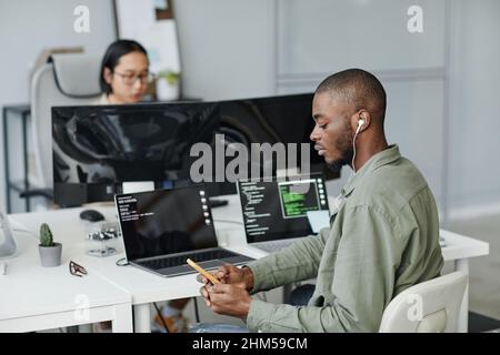 Young African American office worker in earphones scrolling in mobile phone while sitting by desk in front of laptops Stock Photo
