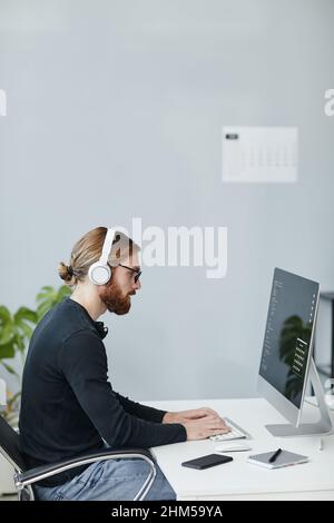 Contemporary young diversity programmer in casual clothes and headphones working in front of computer monitor Stock Photo