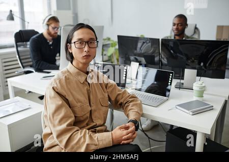 Young serious Asian businessman in eyeglasses and casual clothes sitting by desk with two laptop computers against his colleagues Stock Photo