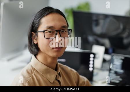 Young Asian businessman in eyeglasses and formal shirt looking at camera while sitting by workplace in contemporary office Stock Photo