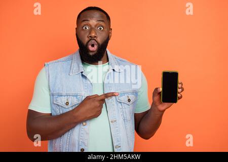 Photo of impressed afro guy wear denim vest pointing modern device empty space isolated orange color background Stock Photo