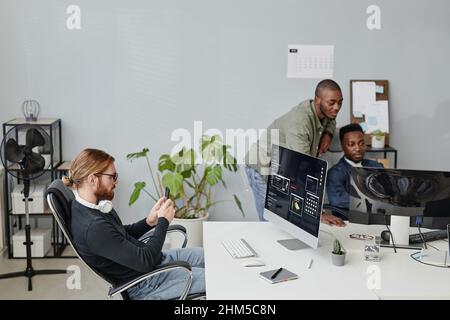Young businessman with smartphone sitting by desk in front of computer screen against two African American coworkers Stock Photo