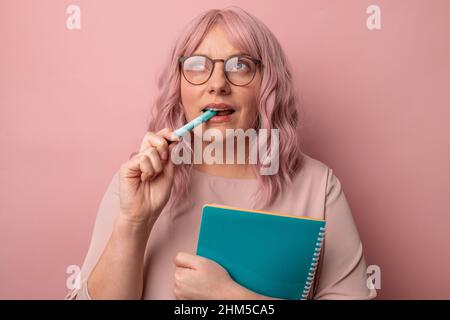 Thoughtful female student wearing glasses holds pen notebook has pensive expression on pink background. Stock Photo