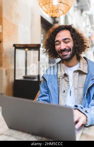 Happy Hispanic man in casual clothes with beard and curly hair smiling. He is using netbook while sitting at table and working on remote project in street cafe Stock Photo