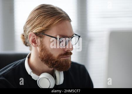 Bearded young man in eyeglasses and black pullover looking at screen of gadget during online communication or distant learning Stock Photo