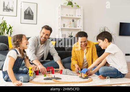 Lovely family of four spend weekend together at home, parents and two kids playing toys, sibling building railway, have a fun. Mom, dad and children sitting on floor in cozy living room and playing Stock Photo