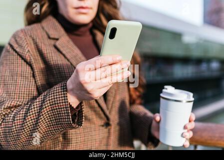 Soft focus of crop woman in checkered coat with eco friendly cup of coffee to go browsing social media on cellphone Stock Photo