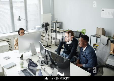 Two young program developers in casual clothes sitting in armchairs by desk in front of computer monitors and working over new program Stock Photo