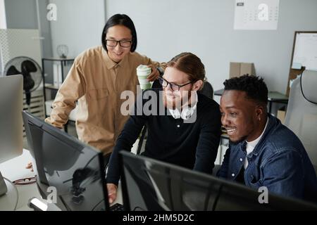 Three cheerful young intercultural diversity programmers decoding data on computer screen at working meeting in bureau Stock Photo