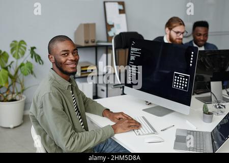 Happy young African American diversity programmer in casual clothes sitting by desk in front of computer monitor with data and icons on screen Stock Photo