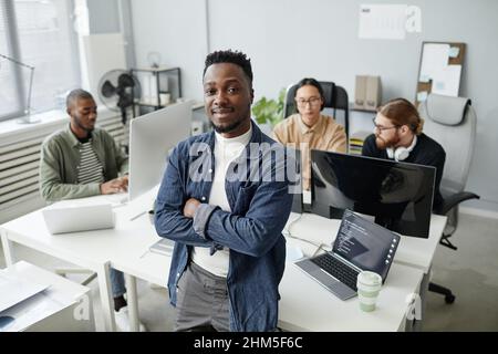 Happy young African American program manager in casual clothes crossing arms on chest while standing against workplace and colleagues Stock Photo