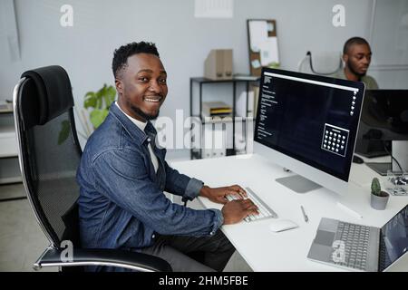 Smiling young African American programmer looking at camera while sitting in armchair by desk and working by computer Stock Photo