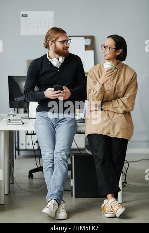 Two young intercultural businessmen in casual clothes standing by desk with computer monitor and discussing their plans for the night Stock Photo