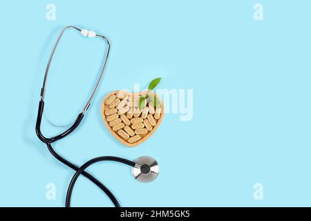 Herbal pills in heart shaped wooden plate and stethoscope on blue background. Alternative medicine Stock Photo