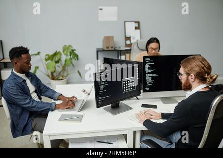 Group of young intercultural software engineers working over new program while sitting by desks in front of computer monitors Stock Photo