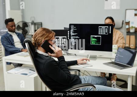 Young busy programmer talking on mobile phone while sitting in front of computer monitor against two intercultural colleagues Stock Photo