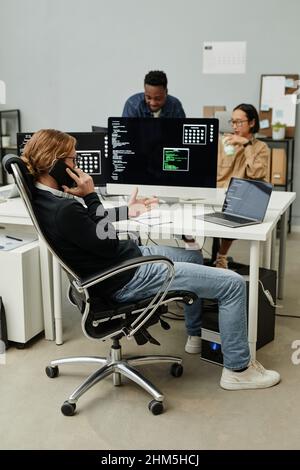 Young Caucasian male programmer calling by mobile phone by workplace while his two colleagues discussing data on computer screen Stock Photo