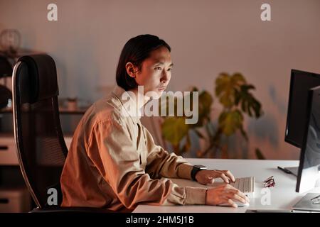 Contemporary young Asian businessman sitting in front of computer monitors while working late in openspace office Stock Photo