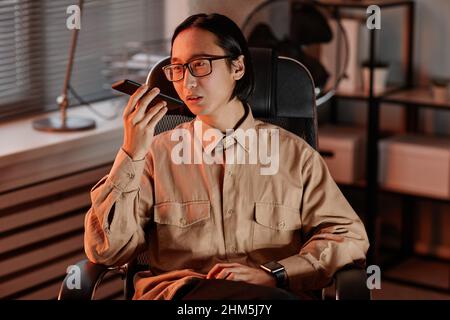 Young Asian businessman in eyeglasses and casual shirt recording voice message while holding mobile phone by his mouth Stock Photo