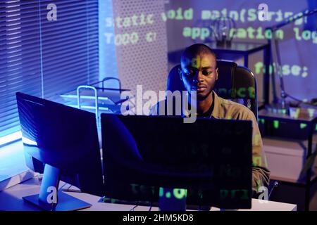 Young serious African American businessman sitting by desk in front of computer monitor while working late at night in office Stock Photo