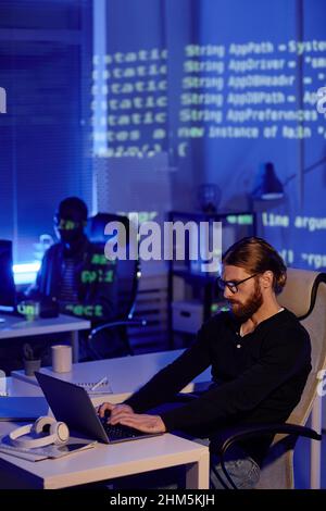 Serious bearded young businessman working in front of laptop while sitting by desk against male colleague in openspace bureau Stock Photo
