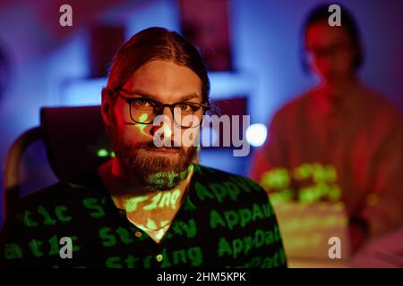 Face of young serious diversity programmer in eyeglasses sitting in front of camera in office late in the night and working Stock Photo