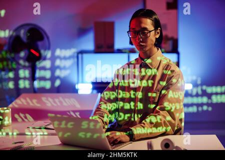 Young Asian male diversity programmer working in front of laptop late at night while sitting by desk in modern openspace office Stock Photo
