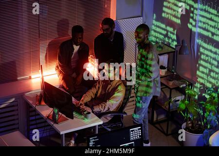 Group of young intercultural software developers standing by colleague typing on computer keyboard while working at night meeting Stock Photo