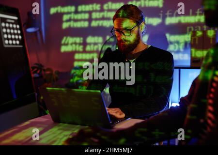Serious bearded software developer looking at data on laptop screen while working over new program late in the night in office Stock Photo