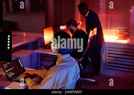 Young Asian software developer working over new program while sitting by desk and looking at laptop screen against two colleagues Stock Photo