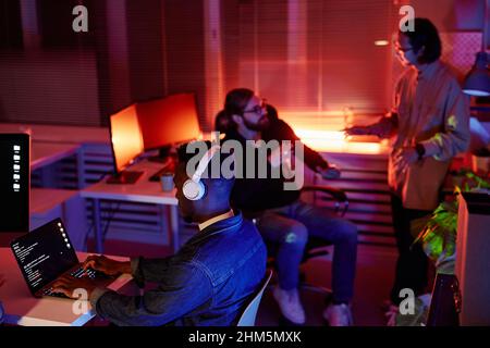 Young male diversity programmer of African ethnicity working in front of laptop while his colleagues having discussion at night meeting Stock Photo