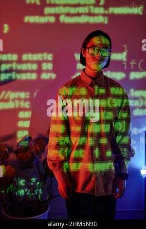 Young serious Asian businessman in casual clothes and eyeglasses standing in openspace office against programming codes Stock Photo