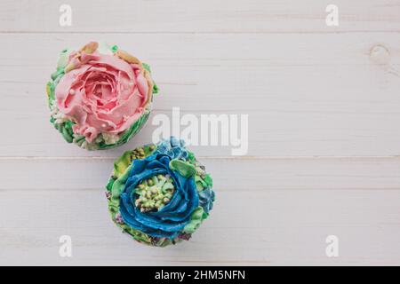 Two muffins with colored butter flowers on white wooden background, flat lay Stock Photo